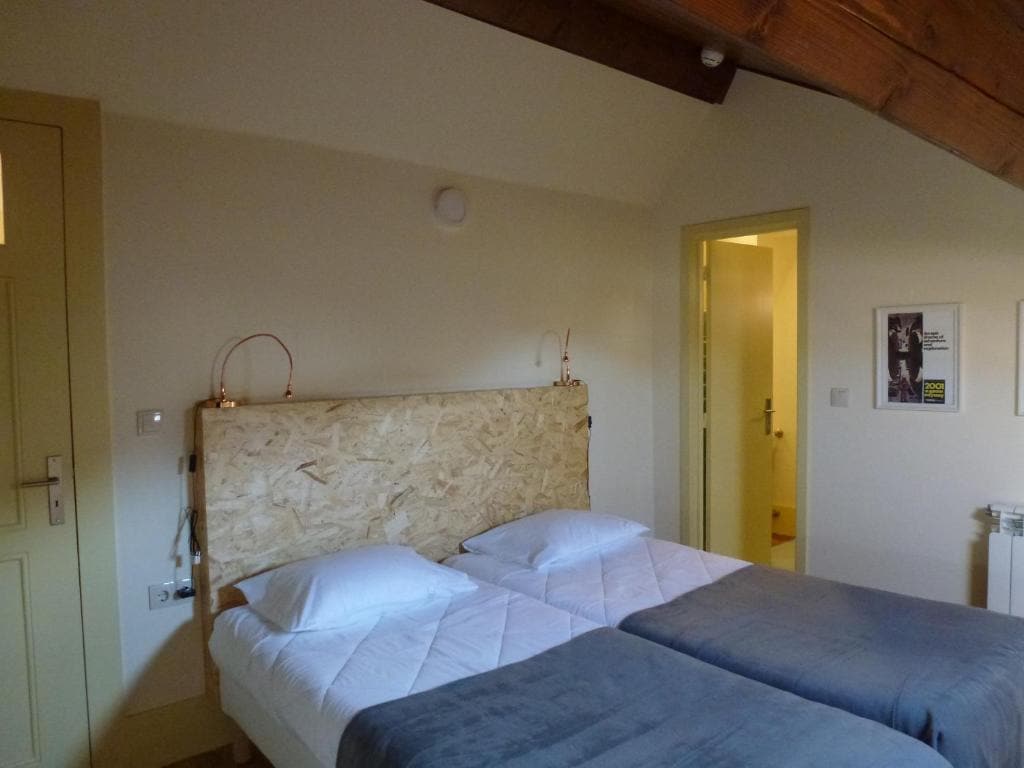 A twin room features two individual beds with crisp white linens and gray bedspreads. A wooden headboard adds a natural element to the space. The room is completed with a door leading to a private bathroom and warm wooden beams visible along the ceiling.