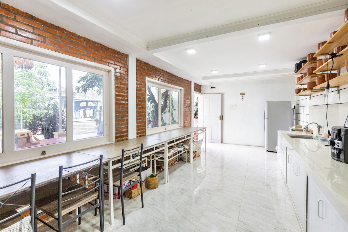 A spacious kitchen area is illuminated by natural light from large windows, offering views of the garden. Brick walls provide a rustic contrast to the modern cabinetry. A long dining table with multiple chairs invites shared meals, while appliances are neatly arranged along the counter.