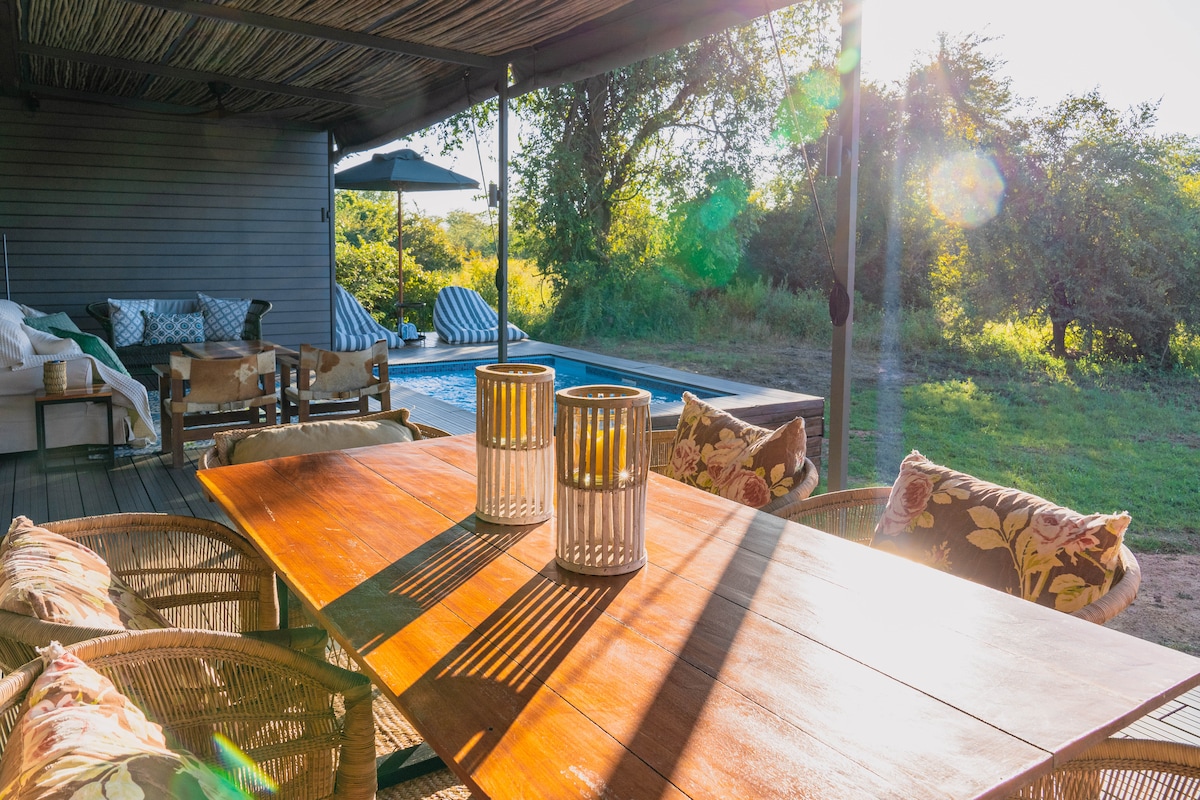 An outdoor dining area is set on a wooden deck, featuring a large table surrounded by chairs with cushions. Soft sunlight filters through the overhead structure, casting gentle shadows. A plunge pool and greenery are visible in the background, enhancing the relaxed atmosphere.