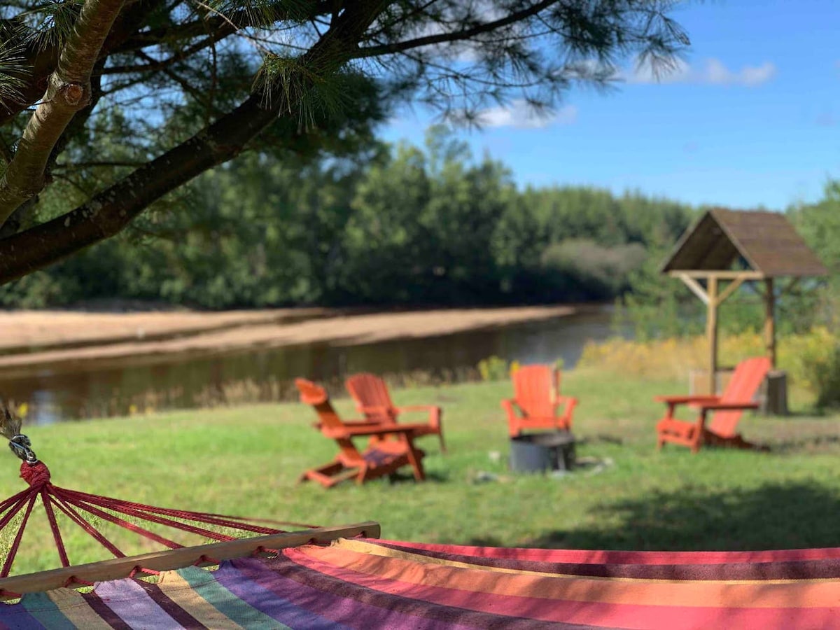 A hammock is gently swaying between trees, overlooking a serene river. Red Adirondack chairs are arranged around a fire pit on a grassy area, with a gazebo in the background, all surrounded by lush greenery and a clear blue sky.