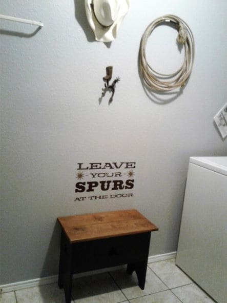 A welcoming entryway features a black wooden bench against a light gray wall. Text stating 'LEAVE YOUR SPURS AT THE DOOR' is displayed in a decorative font. Country-themed decor includes a cowboy hat, rope, and a small decorative hook, lending a charming southwestern feel.