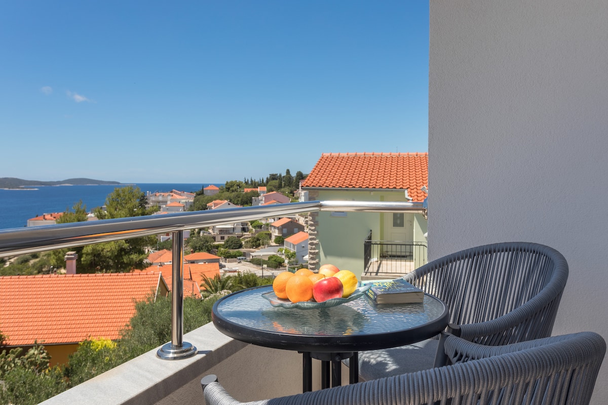A balcony features a glass table set with a variety of colorful fruits. Two woven chairs provide a comfortable seating area, overlooking a scenic view of homes and the coastline. The clear blue sky and the sea beyond enhance the tranquil setting.
