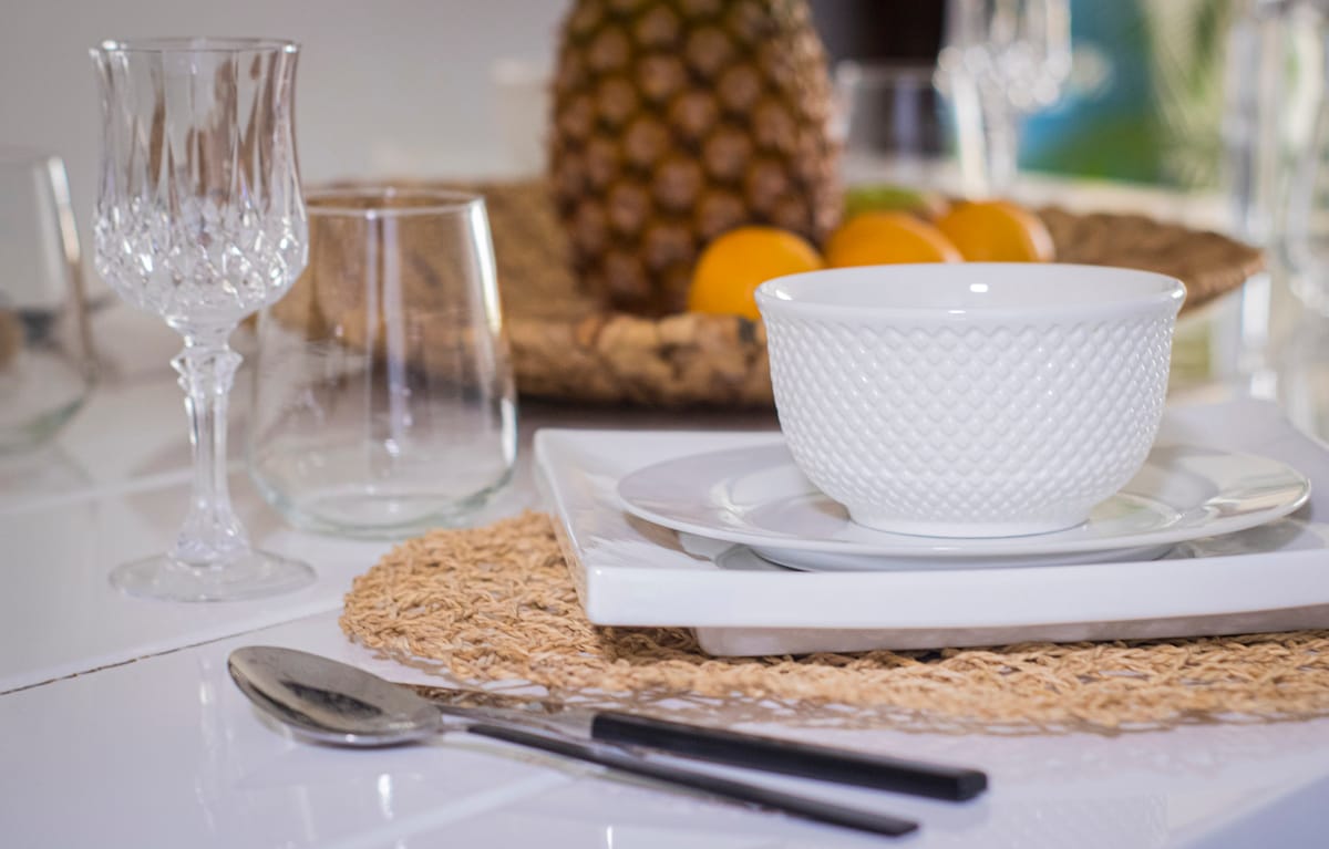 A dining setting is presented with elegant glassware and white dishware arranged on woven placemats. A decorative bowl sits atop a white plate, accompanied by polished cutlery. In the background, a bowl filled with oranges and a pineapple adds a touch of freshness.