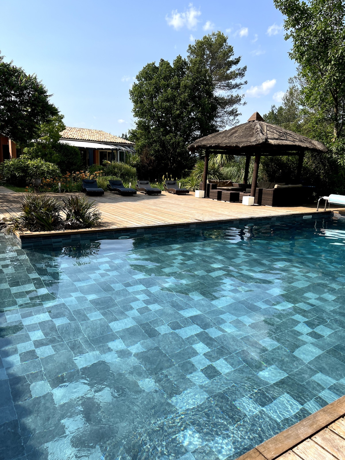 The heated pool reflects the clear blue sky, surrounded by a wooden deck with lounge chairs and a shaded gazebo. Lush greenery and flowering plants frame the area, providing an inviting setting for relaxation and enjoyment.