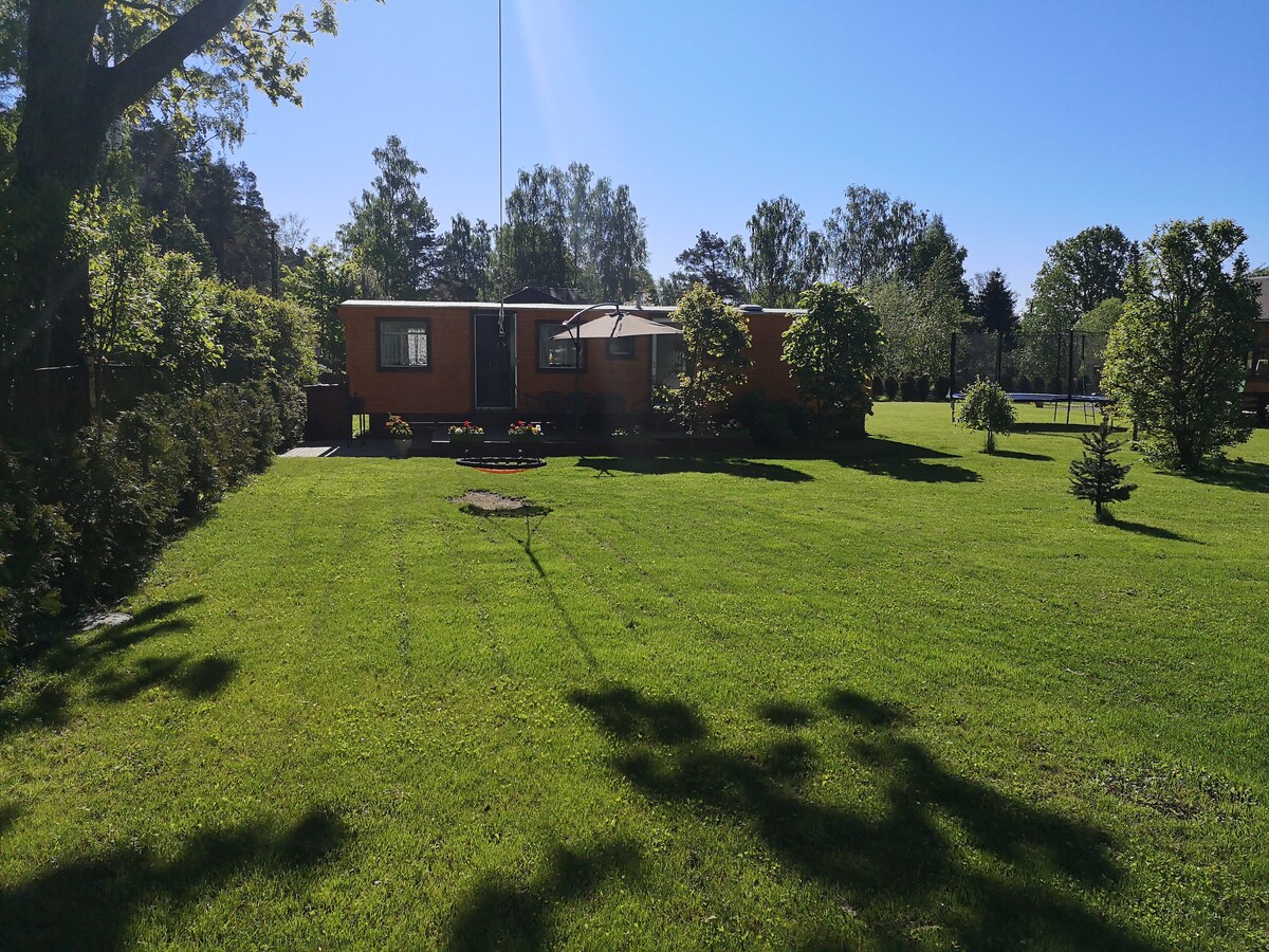 A well-maintained outdoor area is presented with a spacious green lawn, framed by trees and shrubbery. The cozy dwelling stands in the background, featuring a canopy that provides shade. A large trampoline can be seen to one side, contributing to the recreational space.