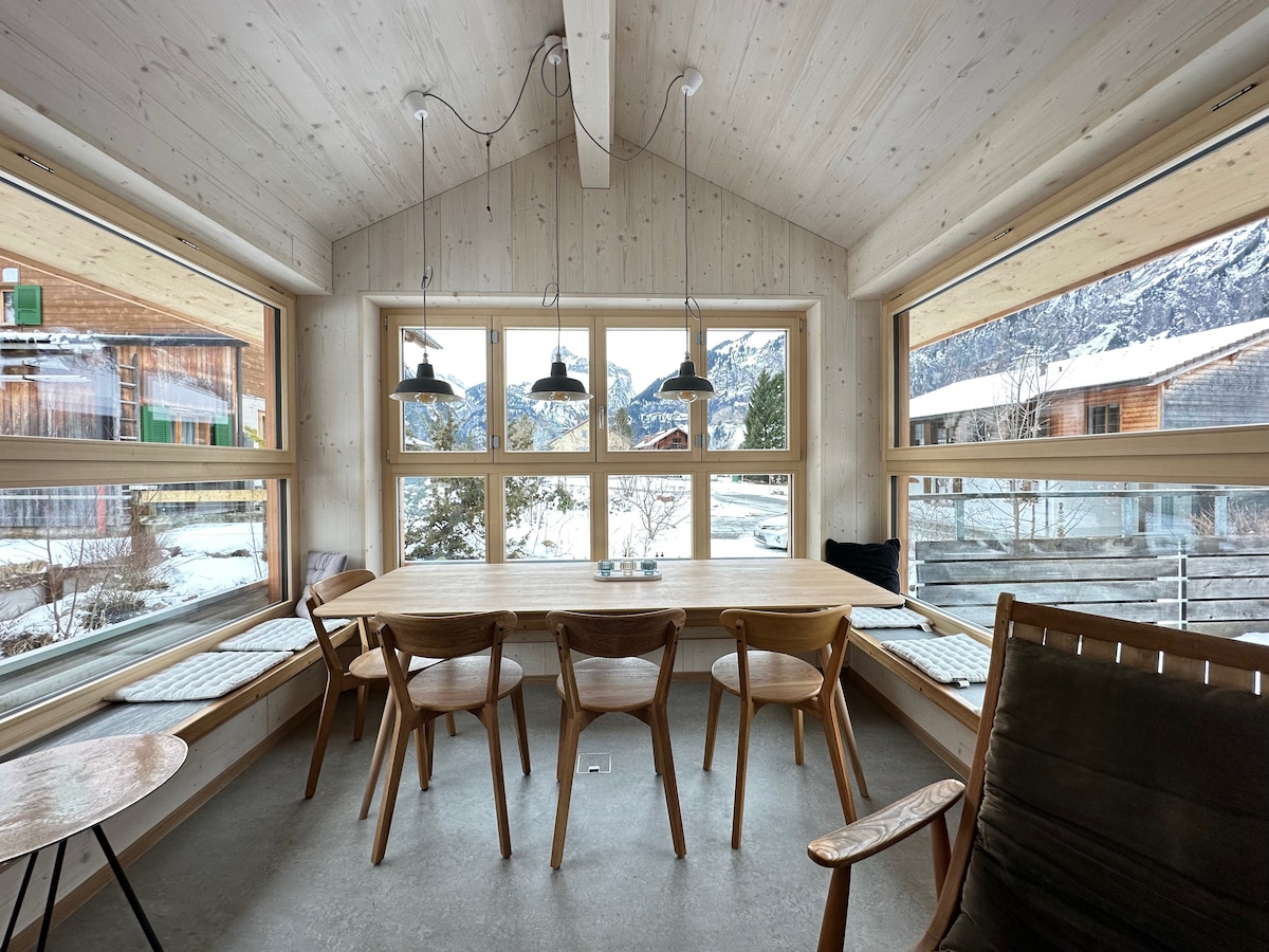 A spacious dining area is presented with a wooden table surrounded by chairs, accentuated by large windows letting in ample natural light. The view includes a winter landscape with snow-covered surroundings and mountain scenery. Two pendant lights add a modern touch to the design.