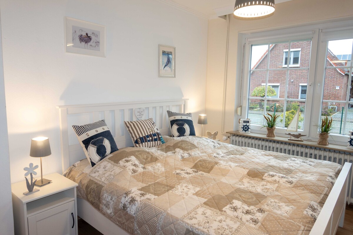 A bright bedroom is presented with a spacious bed, adorned with a patterned quilt. Nightstands on either side hold lamp fixtures, providing soft illumination. Natural light enters through a large window, revealing a view of the surrounding greenery and the exterior of neighboring houses.