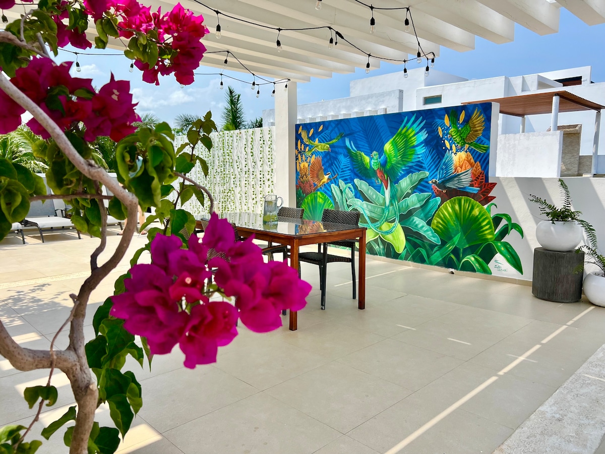 An outdoor dining area is featured beneath a shaded terrace, with a large table and chairs set against a vibrant mural of tropical flora and fauna. Bougainvillea blooms in shades of pink add a natural touch alongside minimalist white planters, contributing to the serene ambiance.
