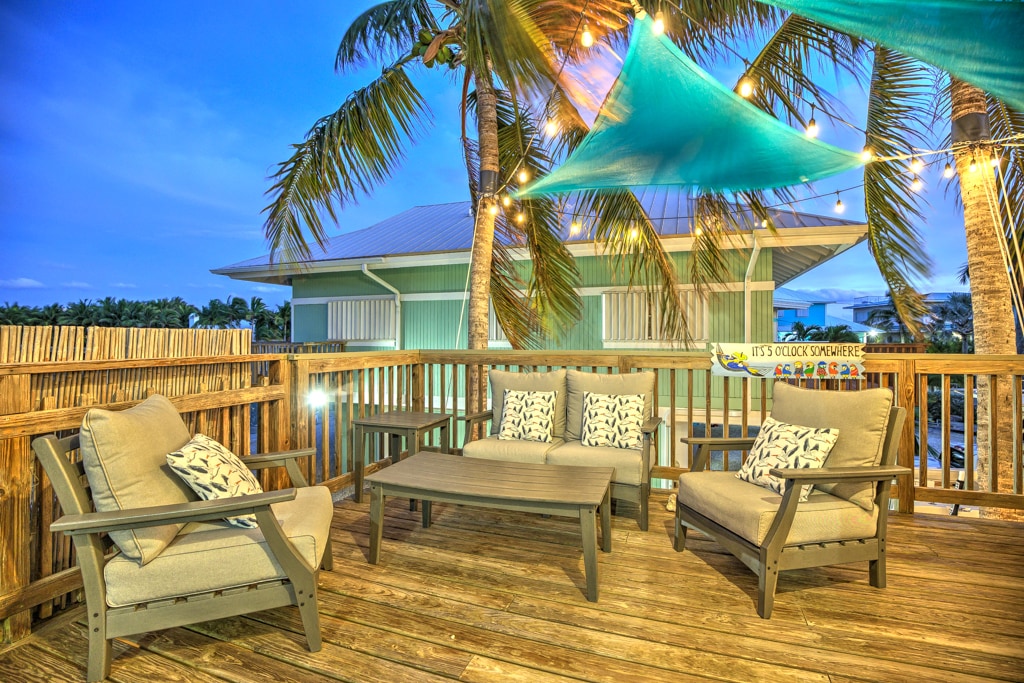 An outdoor deck space is featured with comfortable seating arrangements, including two lounge chairs and a central table. Decorative pillows add a pop of color. String lights are draped overhead, and palm trees provide shade, enhancing the relaxed environment suitable for gatherings.