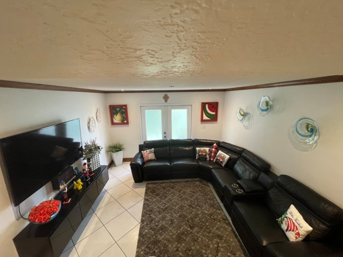 The living area features a spacious sectional sofa arranged around a television. Decorative wall art in vibrant colors adds interest to the surroundings. Large doors provide access to an outdoor space, while a few potted plants complement the room's layout.