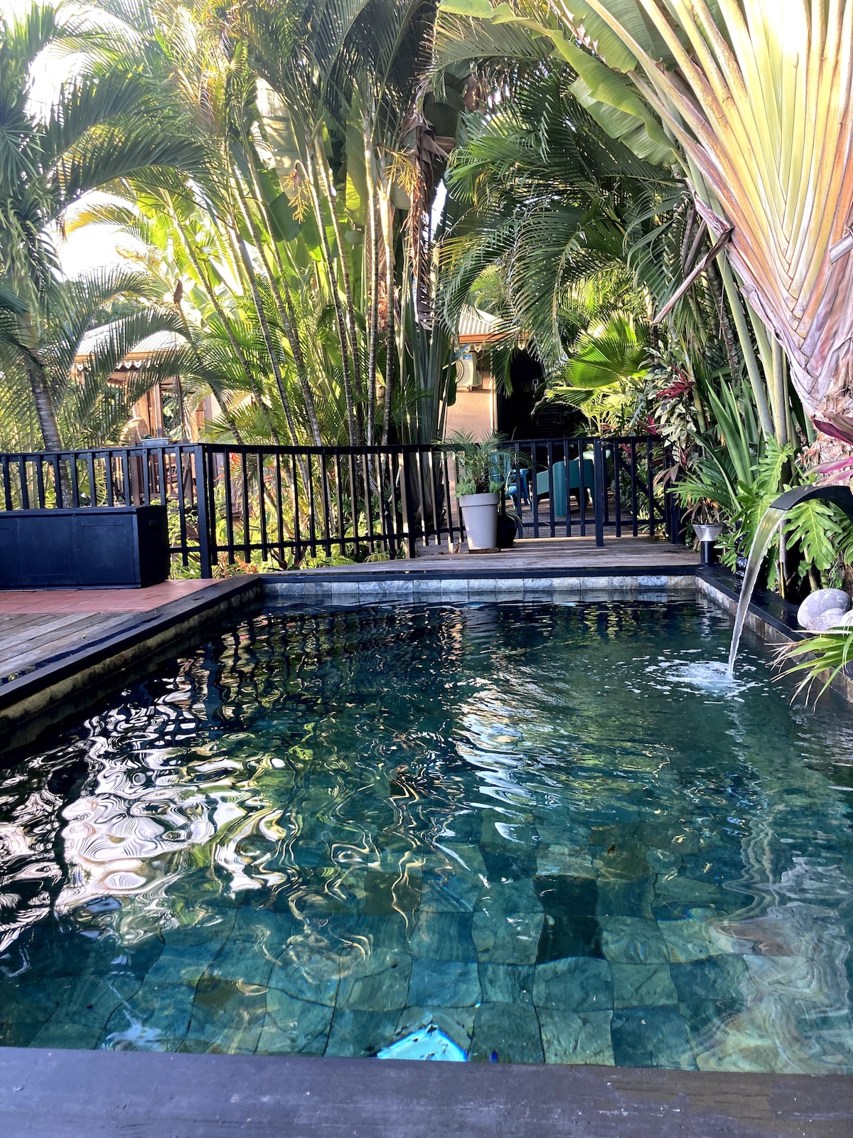 A private pool is surrounded by lush tropical vegetation, with tall palm trees and vibrant plants creating a serene atmosphere. The water reflects the natural light, showcasing its clear, inviting surface. A wooden deck complements the pool area, ideal for relaxation.