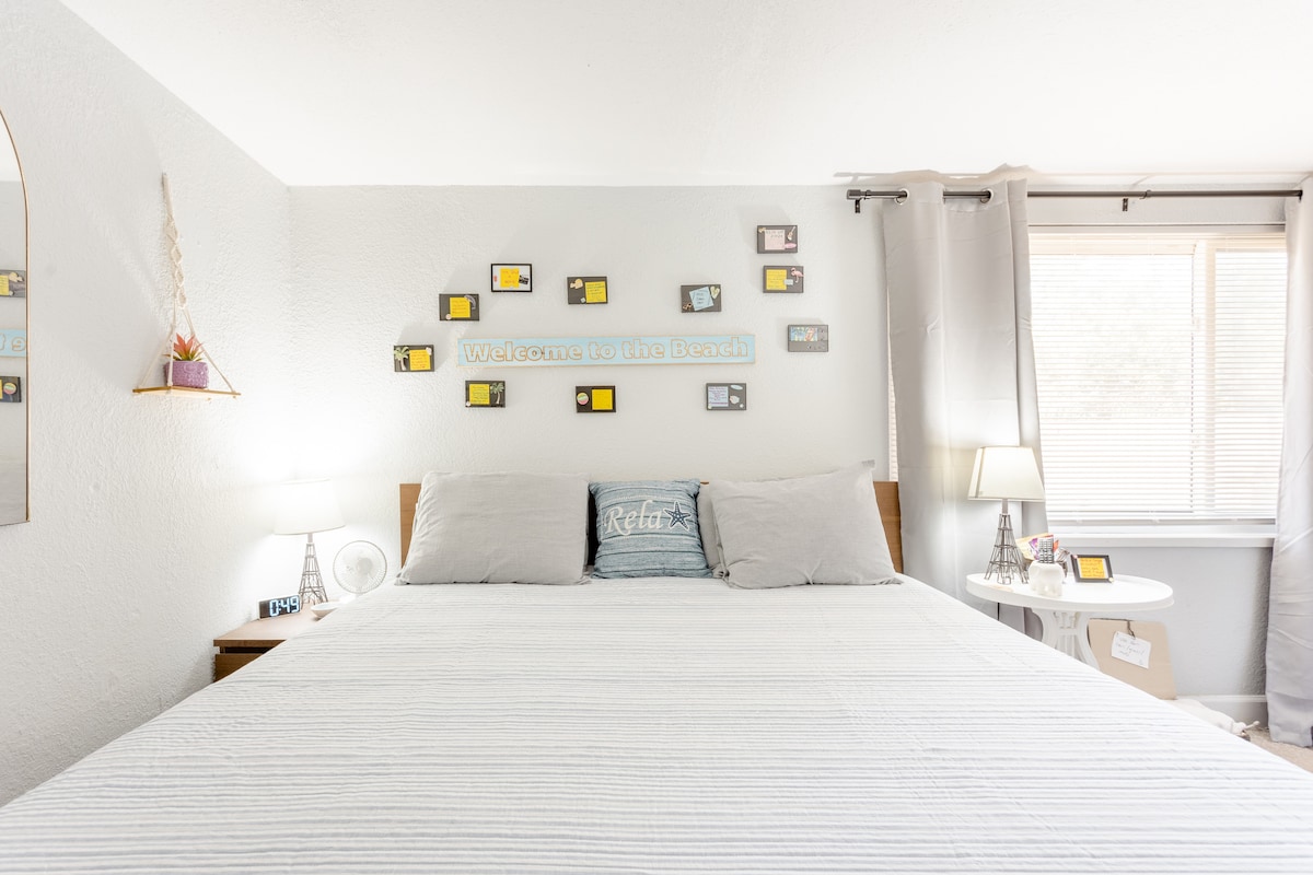 A spacious bedroom features a king bed with light bedding and decorative pillows. The wall displays several framed images and a welcome sign. Soft lighting is provided by bedside lamps, and natural light enters through a nearby window with sheer curtains.
