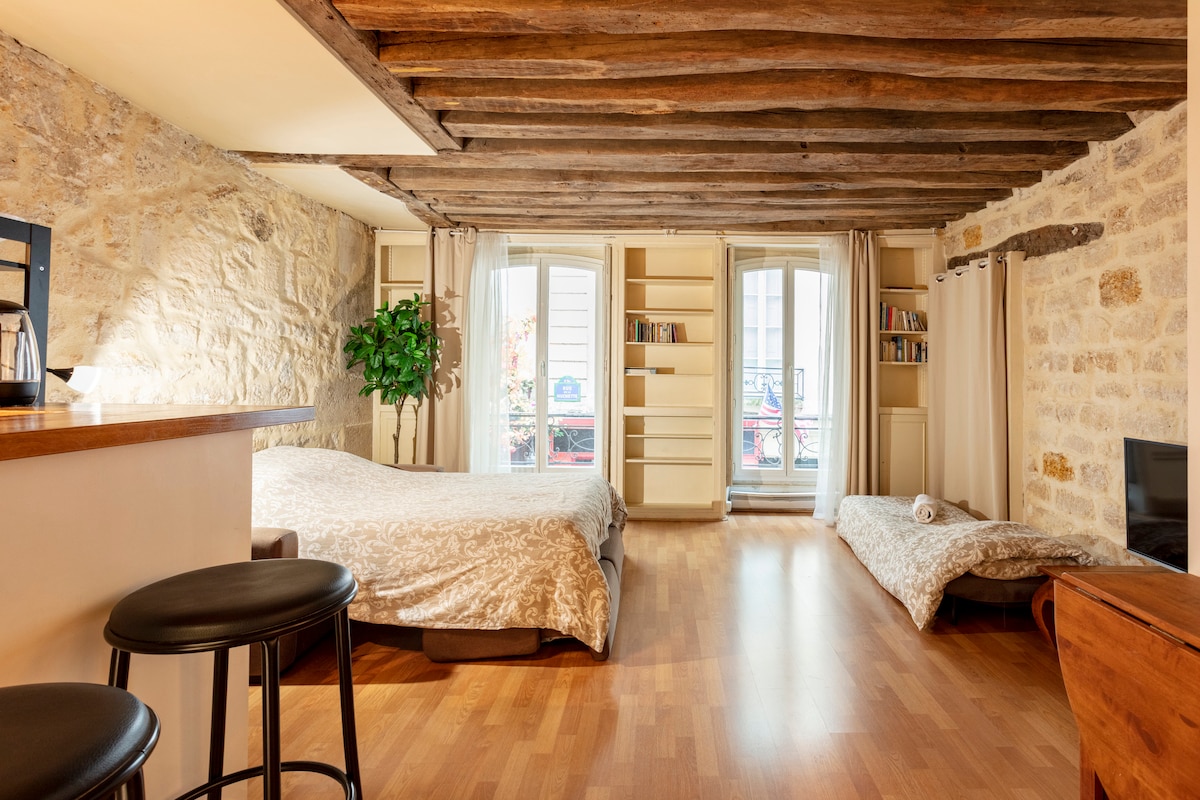 A spacious room features exposed wooden beams and natural stone walls, creating a blend of modernity and charm. Two sleeping areas include a larger bed and a single sofa bed. A small kitchenette is visible, and light enters through double doors leading to a balcony.