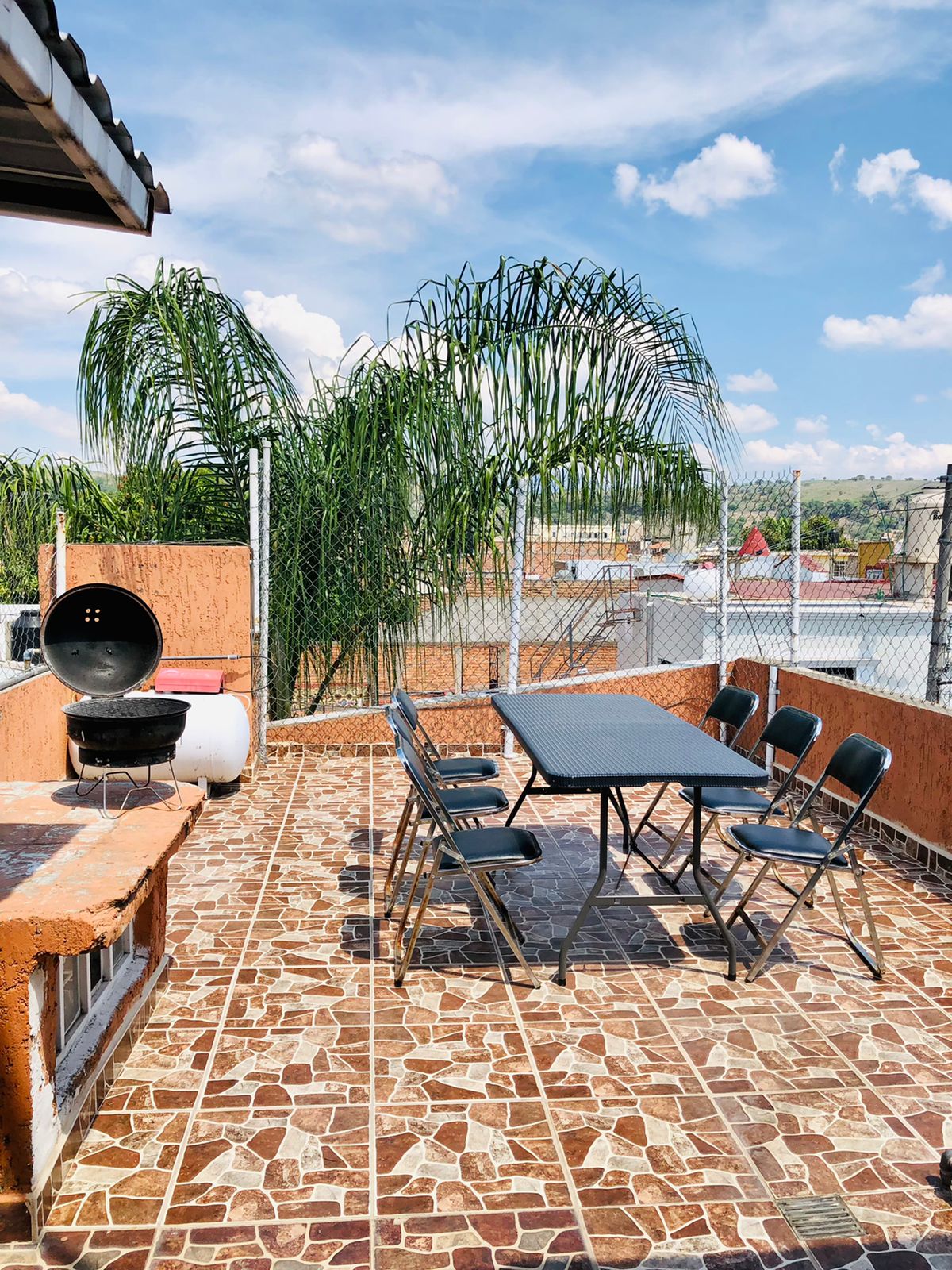 A rooftop terrace features a dining table accompanied by six black chairs. A barbecue grill is positioned nearby. Surrounding greenery and a chain-link fence offer privacy while enhancing the outdoor experience. The floor is tiled with a decorative pattern, and bright clouds fill the sky.