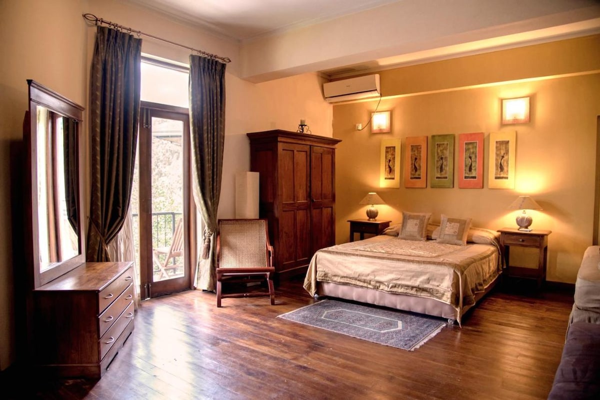 A spacious bedroom features a large bed draped in soft linens, complemented by warm wooden furnishings. Natural light enters through a balcony door, illuminating the rich wood flooring. Artwork is displayed on the walls, providing a touch of color to the serene environment.
