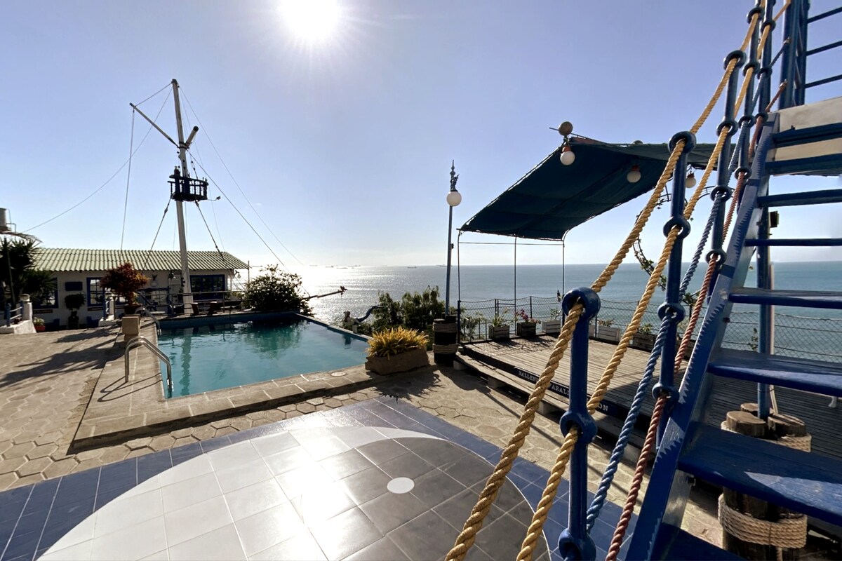 An infinity pool is framed by sunny outdoor spaces, with ocean views extending beyond. A shaded seating area is positioned nearby, while a staircase leads down to the pool. Natural light enhances the inviting atmosphere, highlighting the nearby coastal landscape.