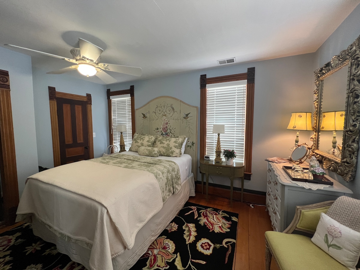 A cozy bedroom is furnished with a spacious bed featuring a floral headboard and soft, neutral bedding. Natural light enters through two windows, while a decorative mirror and a side table with lamps add warmth. A colorful area rug enhances the floor space.