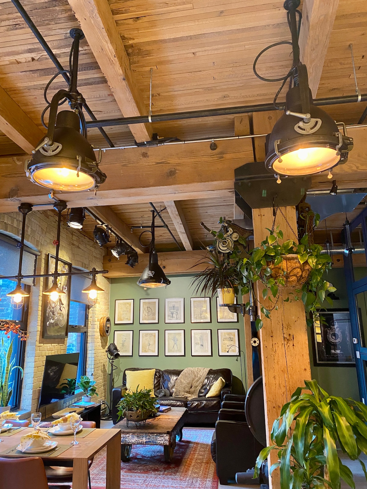 An industrial-style living area features exposed wooden beams and brick walls, with vintage-style lighting hanging from the ceiling. A comfortable seating area is adorned with dark leather couches, surrounded by green plants. A dining table set for a meal adds to the inviting layout.