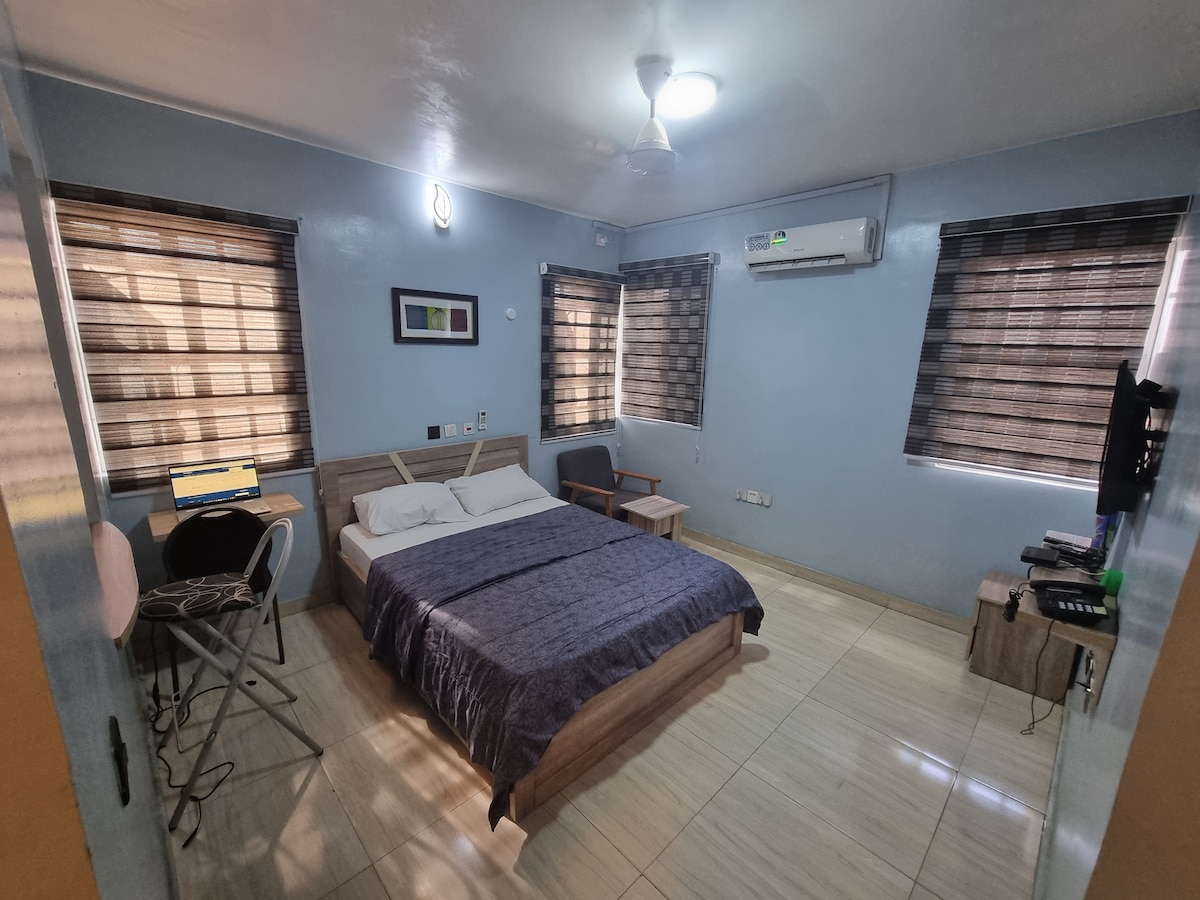 A well-furnished room is presented with a spacious bed covered by a dark blue blanket. Two windows with striped blinds allow natural light in. A small desk with a chair and a wall-mounted television are visible, alongside an air conditioning unit for comfort.