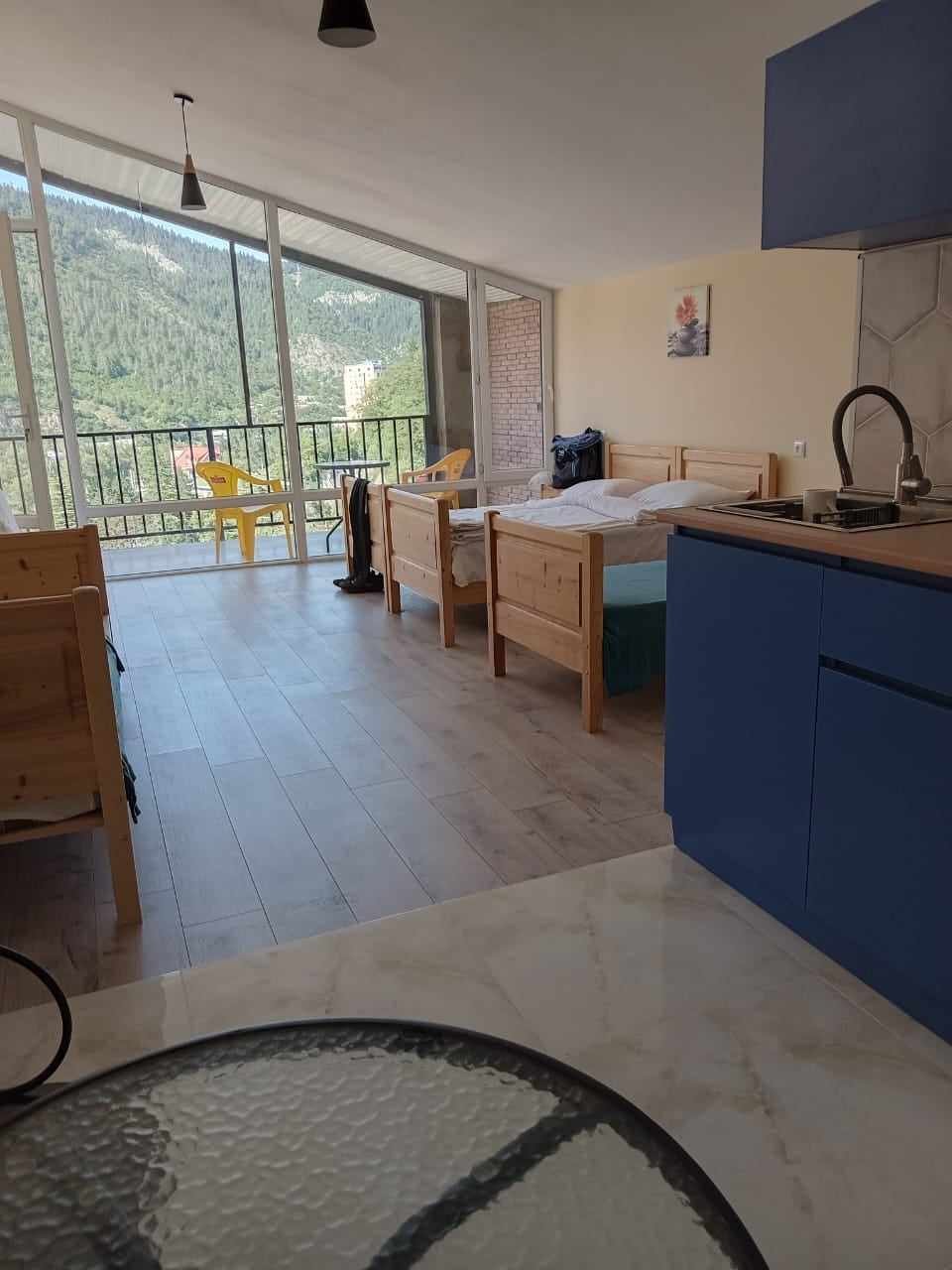 The interior space features wooden furnishings, including two beds and a small kitchen area with blue cabinetry. Large glass doors provide natural light and views of the surrounding mountains. A balcony can be seen, enhancing the connection with the outdoors.
