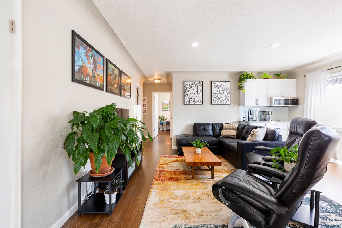 A bright living room is highlighted by comfortable leather seating and a coffee table made of wood. Green plants add a touch of nature, while artwork adorns the walls. Natural light fills the space, creating a welcoming ambiance.