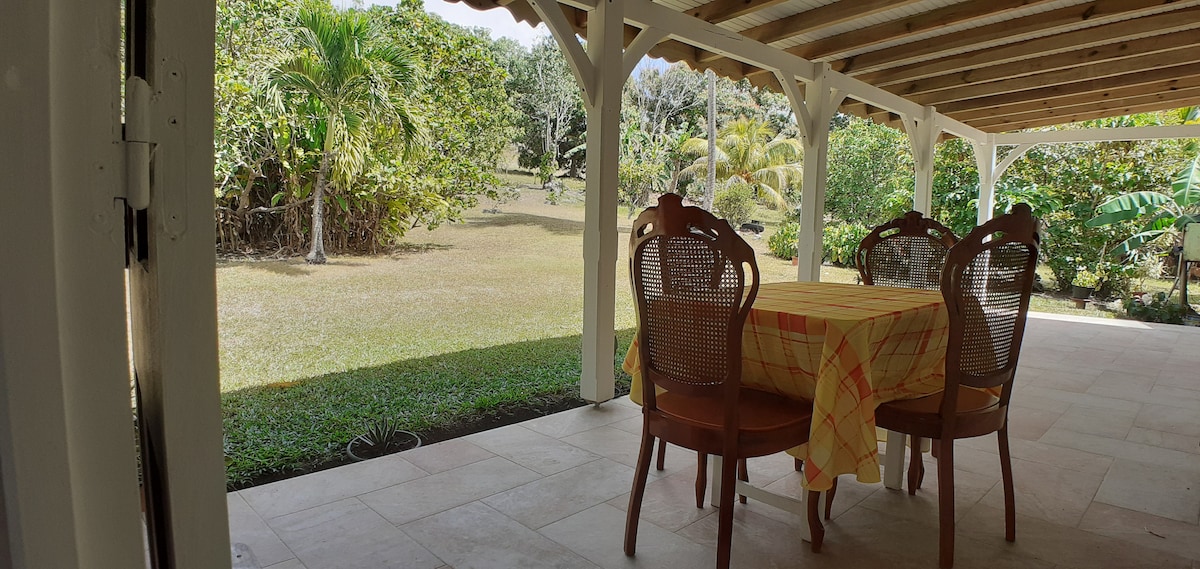 A spacious outdoor dining area is featured under a covered patio, with a brightly colored tablecloth on a round table surrounded by four wooden chairs. Lush greenery and trees provide a natural backdrop, creating a serene atmosphere.