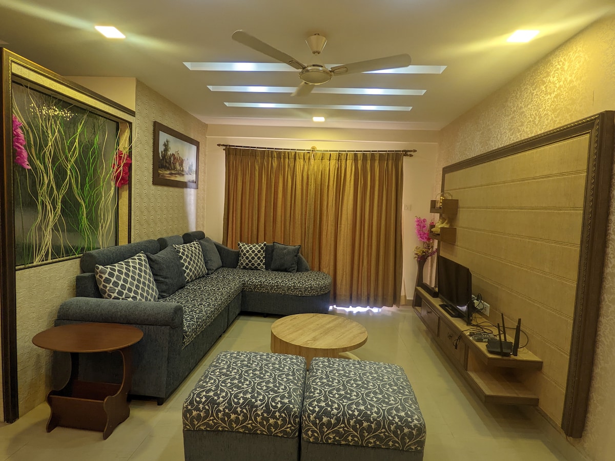The living room features a spacious sectional sofa adorned with patterned cushions, complemented by wooden furnishings. A round table is centrally positioned, while a television is mounted on the wall. Natural light filters through the partially covered windows, enhancing the contemporary decor.