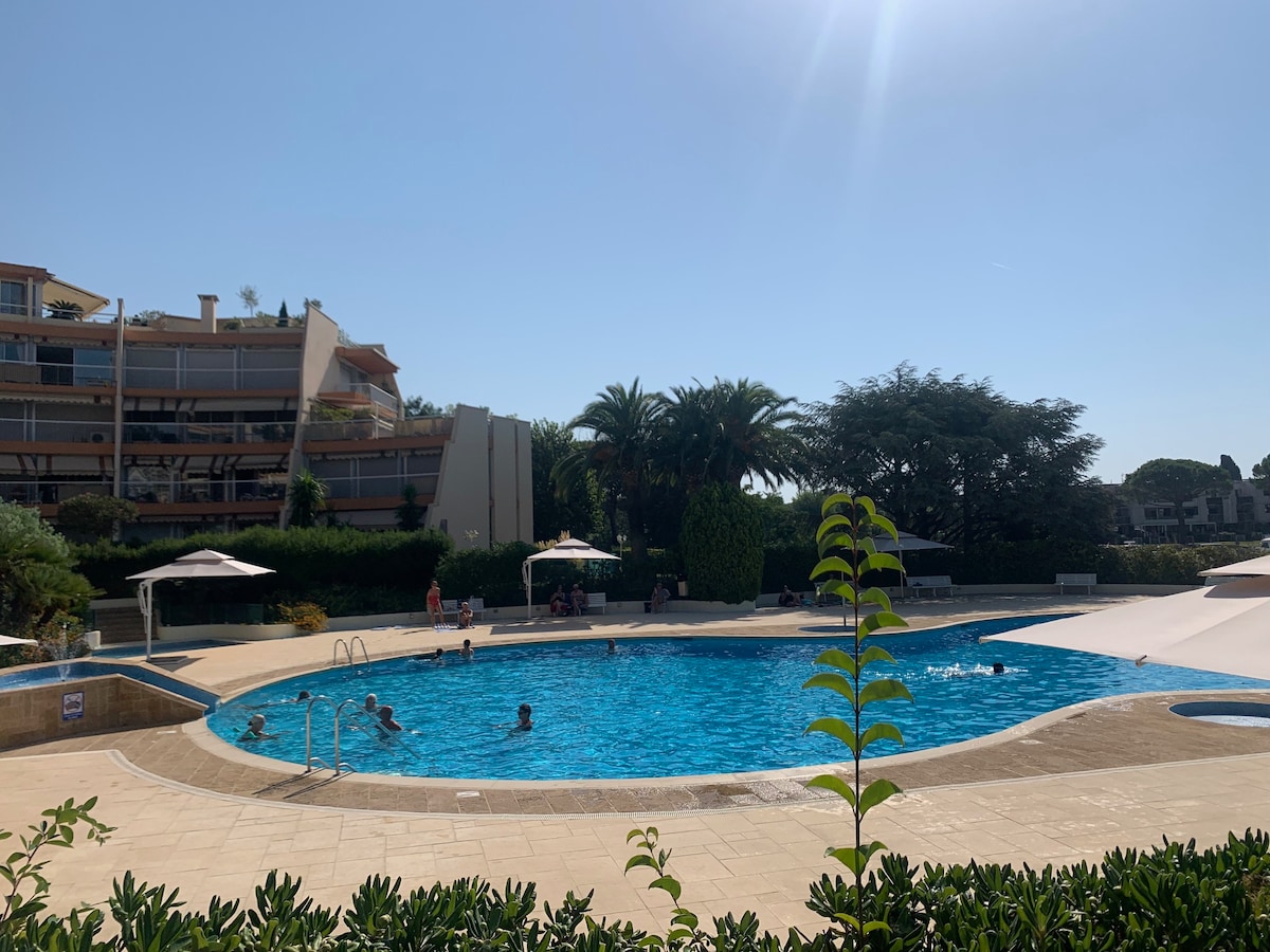A large swimming pool is surrounded by a sun deck with umbrellas and palm trees in the background. The water appears clear and inviting, with several individuals enjoying the space. Lush greenery frames the pool area, providing a sense of tranquility.