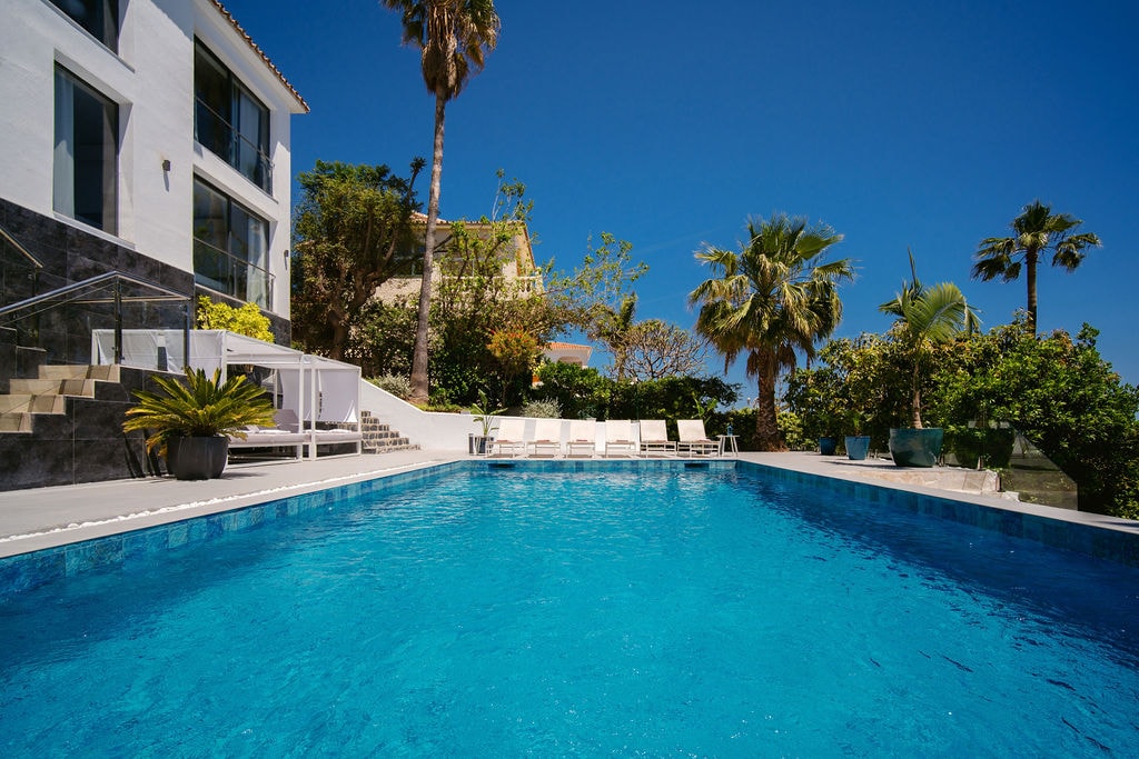 A large heated swimming pool is surrounded by a spacious terrace and lush greenery. Comfortable loungers are positioned nearby under clear blue skies, offering a tranquil setting. Palm trees and various plants enhance the outdoor ambiance, providing a relaxing atmosphere for guests.