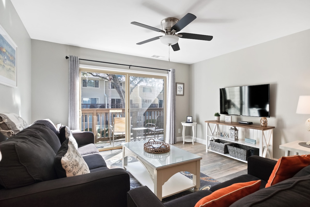 A comfortable living area is highlighted by two dark sofas and a light coffee table. Natural light flows in through sliding glass doors, leading to a small balcony. A ceiling fan is positioned above, and a flat-screen TV is mounted on the wall.