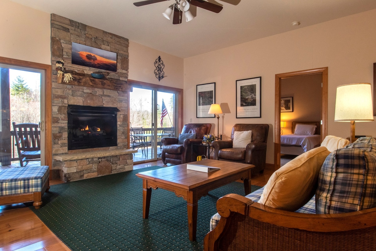 A cozy living area features a stone fireplace as its focal point, surrounded by comfortable leather chairs and a wooden coffee table. Large glass doors provide access to an outdoor deck, allowing natural light to fill the space. Artwork and photos adorn the walls.