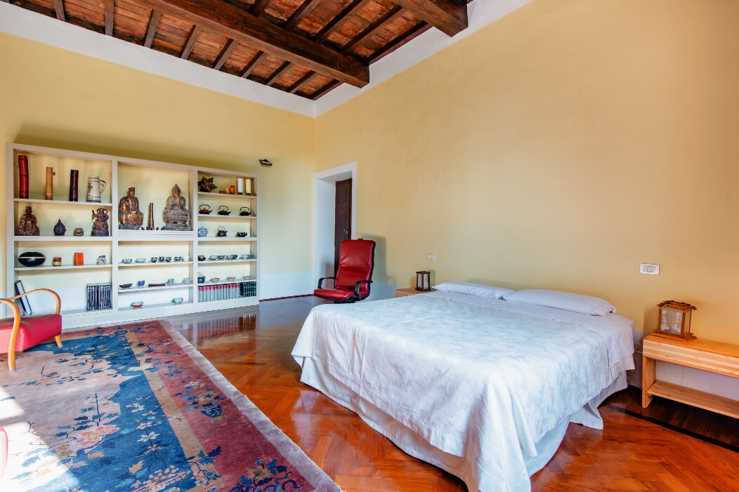 A spacious room features high wooden beams and warm yellow walls. A large, unmade bed is centrally positioned, accompanied by a red recliner. A bookshelf displaying decorative items and books is seen against one wall, while a patterned rug adds texture to the wooden floor.