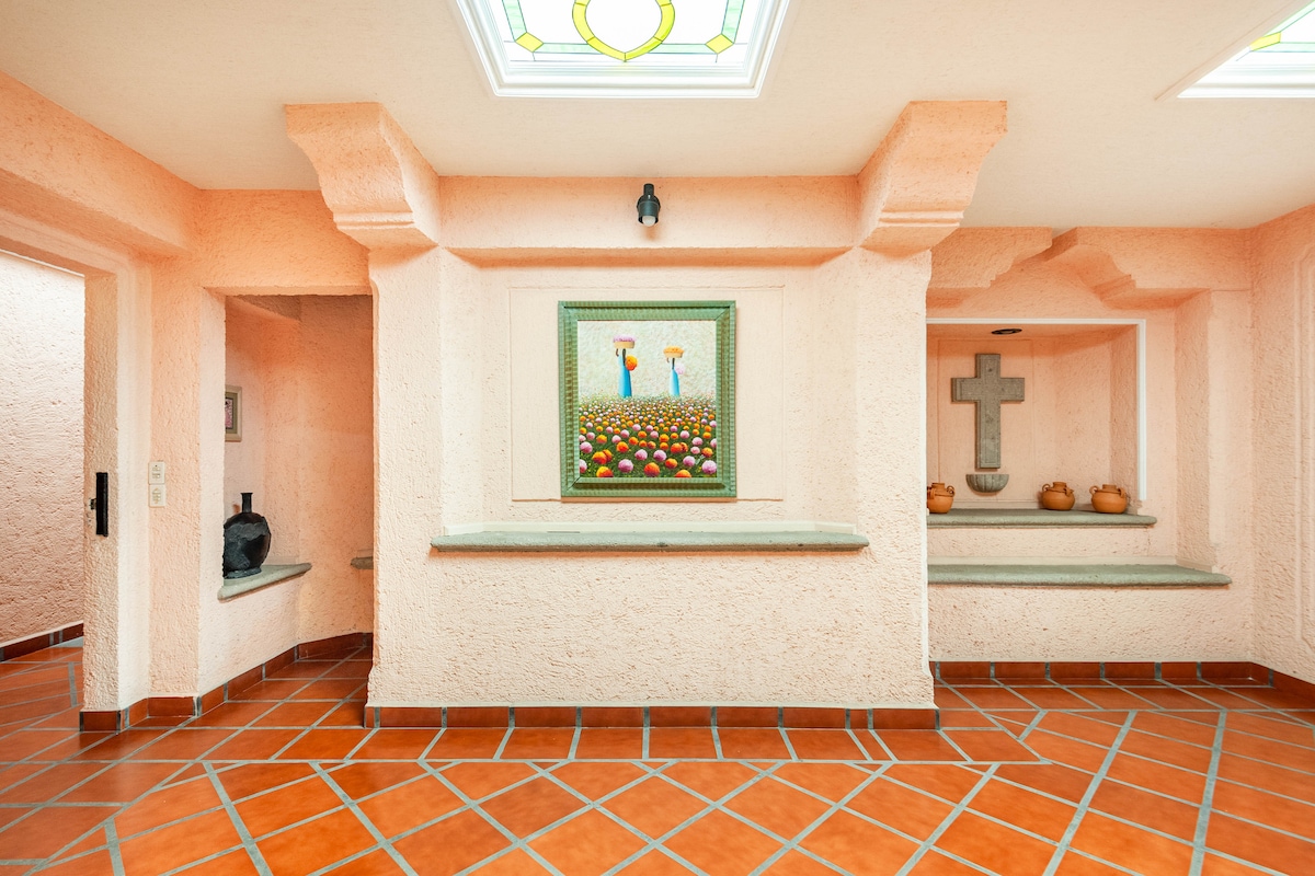 A spacious entryway features textured peach-colored walls and decorative tile flooring. An artwork depicting vibrant flowers is mounted on the wall, while decorative elements are displayed on ledges. Natural light filters through skylights above, enhancing the welcoming ambiance of the space.