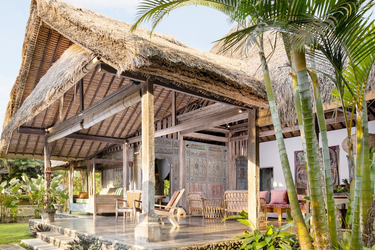 A traditional joglo features a thatched roof supported by wooden beams, surrounded by lush greenery. Open spaces invite natural light, revealing intricate carvings on the walls and comfortable seating areas. The design connects the interior to the tranquil outdoor environment.