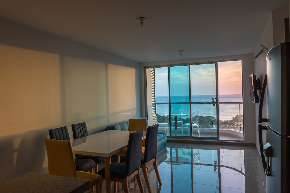 A dining area with a white table and a mix of yellow and blue chairs is seen next to large glass doors. The ocean view is visible, providing a backdrop of soft colors from the sunset. A kitchenette and a television are also present in the space.