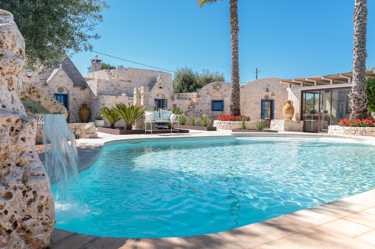 An irregularly shaped pool reflects clear blue skies, bordered by palm trees and vibrant flower beds. A subtle waterfall creates a peaceful ambiance, while a seating area invites relaxation. Trulli-style homes in the background enhance the serene atmosphere of the outdoor space.