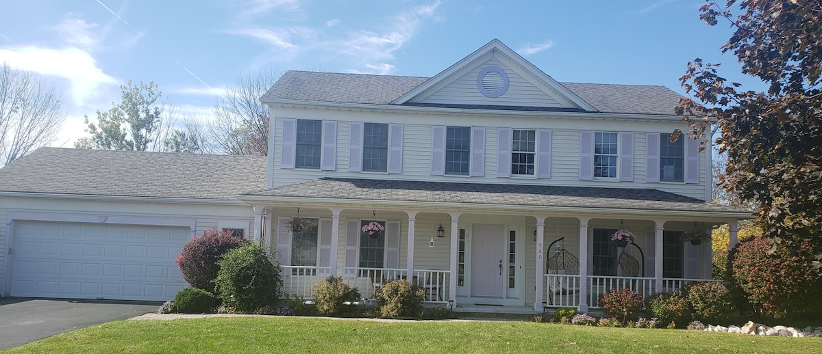 A two-story, suburban home with a light exterior features a welcoming front porch adorned with hanging flower baskets. Green shrubbery and small rocks outline the porch, while a garage is visible on the left. Clear blue skies complement the tranquil atmosphere.