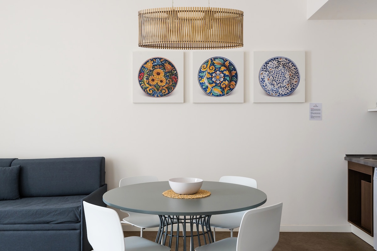 A dining area is presented with a round table surrounded by four white chairs. Above the table, three patterned plates are displayed on the wall, complemented by a woven pendant light. A dark sofa is positioned nearby, providing additional seating.