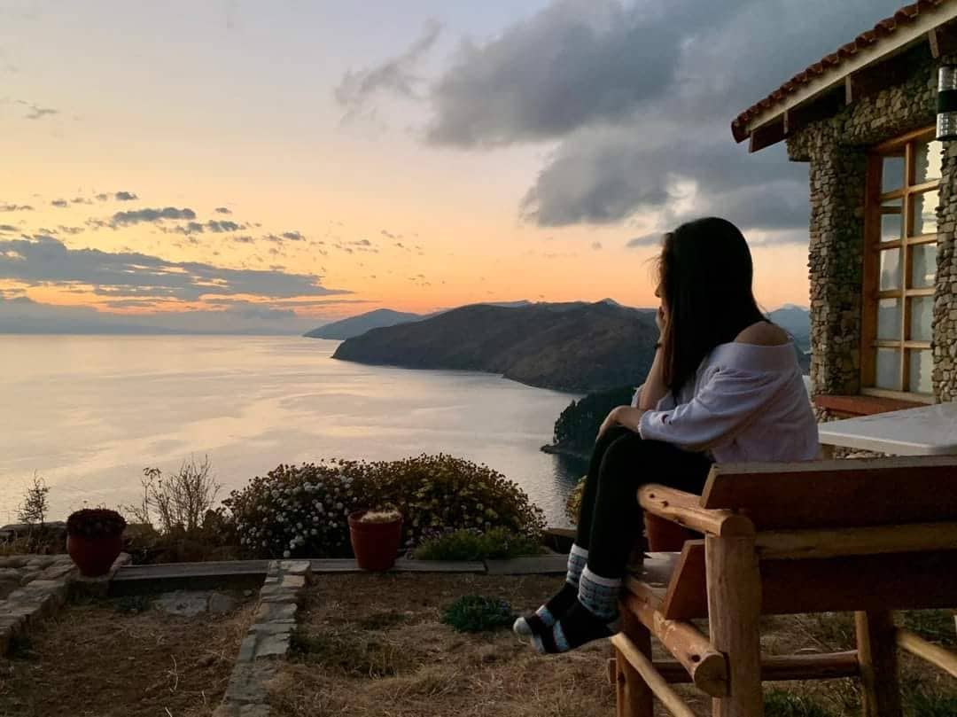 A woman is seated on a rustic wooden chair, overlooking a breathtaking view of a lake at sunset. The landscape features mountains in the distance, with soft clouds scattered across the sky. Nearby, flowers are arranged in pots, adding a touch of nature to the serene setting.