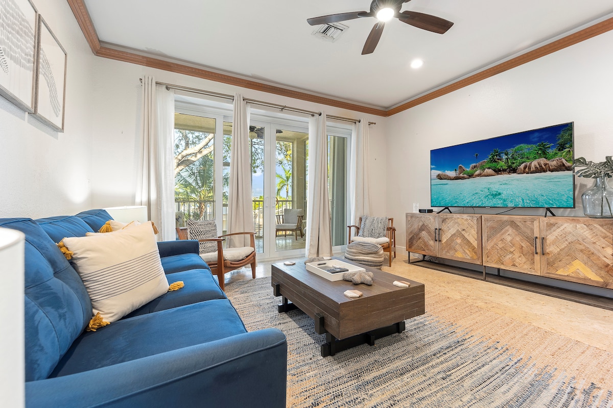 A spacious living area features a blue sofa and a wooden coffee table, complemented by neutral decor. Large glass doors reveal a balcony with views of the outdoors. A flat-screen television is mounted on a stylish console, creating an inviting space for relaxation.