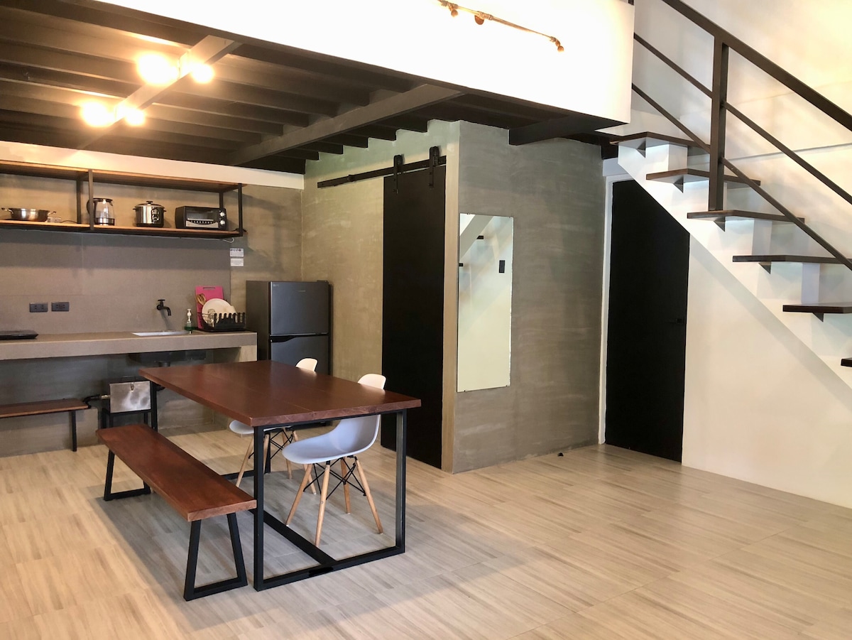An open-plan kitchen and dining area features a wooden dining table with bench seating. A black fridge is placed against a concrete wall, while shelves hold kitchen essentials. Stairs ascend to the second floor, and an accent mirror is mounted nearby.