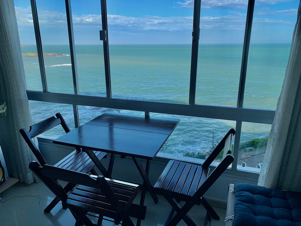 A small wooden dining table and two folding chairs are positioned by large glass windows offering a panoramic view of the ocean. The blue waters stretch towards the horizon, with gentle waves visible beneath a partially cloudy sky.