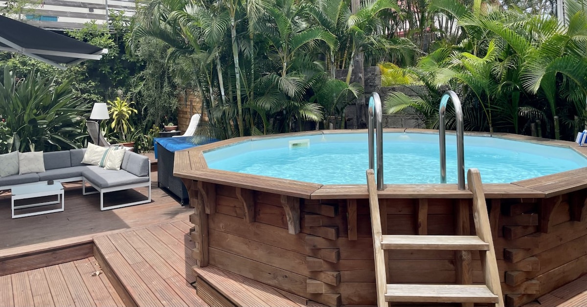 An octagonal wooden pool is surrounded by lush tropical plants and seating areas. Grey outdoor furniture is arranged nearby, providing a comfortable space for relaxation. Sunlight reflects off the clear water, inviting guests to enjoy the outdoor setting.