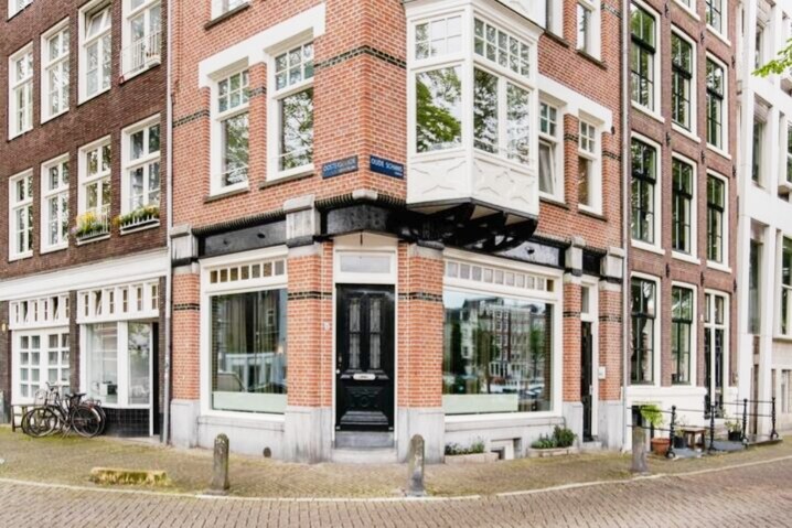 A charming corner building is showcased, featuring large windows that provide ample natural light. The brick facade is contrasted by white trim and a decorative balcony. Bicycles are parked nearby, and the street features a mix of greenery and historic architecture.
