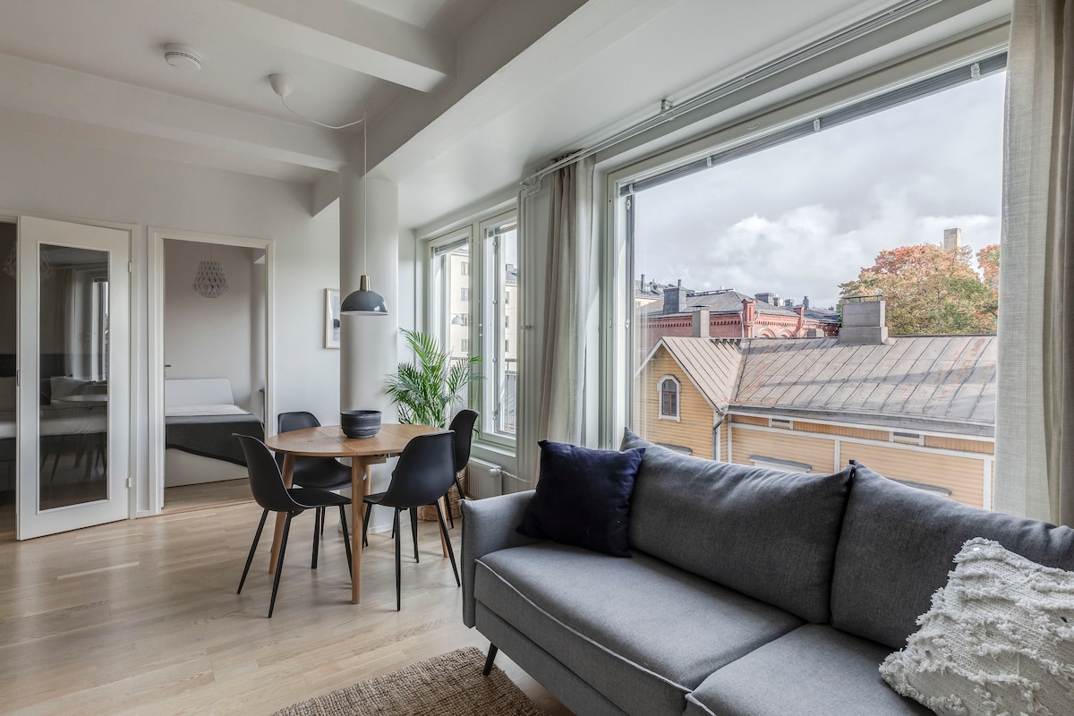 A spacious living area features large windows that provide natural light and a view of the neighborhood. A modern sofa is positioned near a dining table with four chairs. The open layout also allows visibility of another room furnished with a bed.