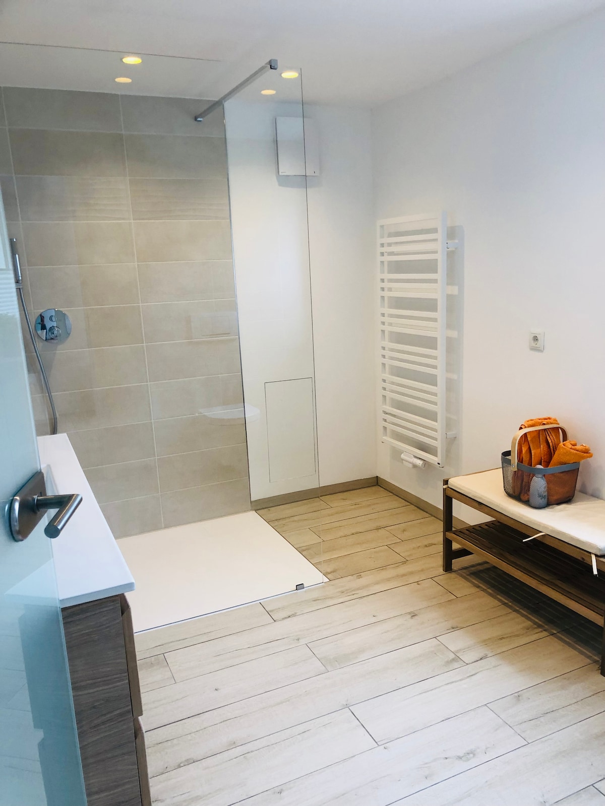 A modern bathroom is presented, featuring a spacious walk-in shower with a clear glass partition. A towel warmer is mounted on the wall, and a wooden bench rests beside a sleek vanity. Neutral tile hues complement the warm wood floor.
