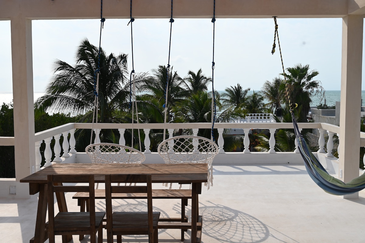 A spacious rooftop terrace features a wooden dining table accompanied by two hanging chairs, set against a backdrop of palm trees and the ocean in the distance. A hammock adds a cozy element, inviting relaxation and enjoyment of the surrounding scenery.