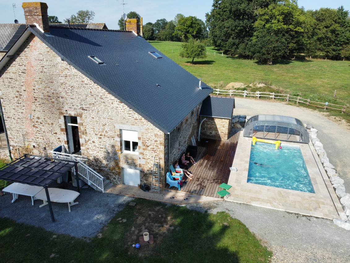 An aerial view displays a charming renovated stone house with a dark roof, accompanied by a fenced pool area and a spacious terrace. Sun loungers are positioned next to the pool, surrounded by rolling green fields and mature trees, creating an inviting outdoor space.