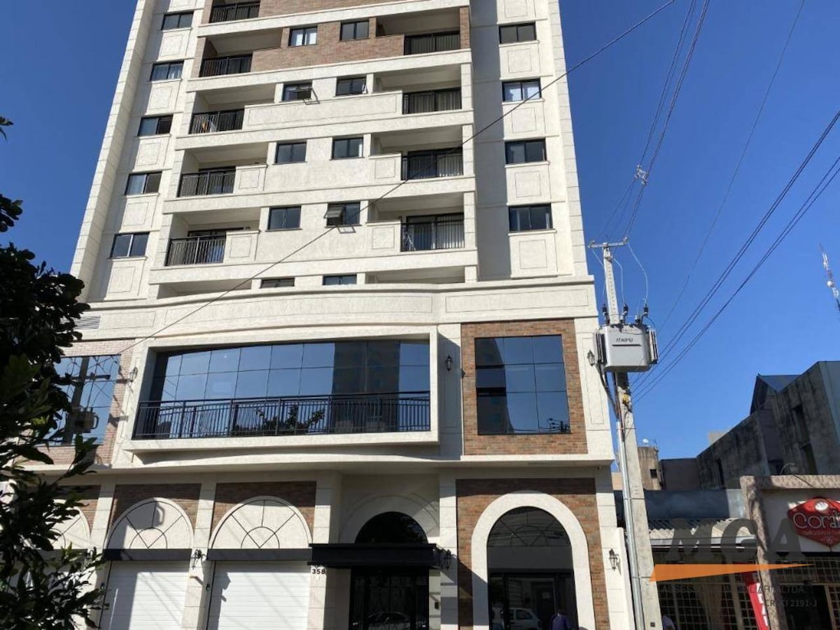 A modern building featuring a light-colored facade with decorative elements. Large glass windows are observed on the ground level, providing a view of the entrance area. The structure is surrounded by urban scenery, with clear blue skies above.
