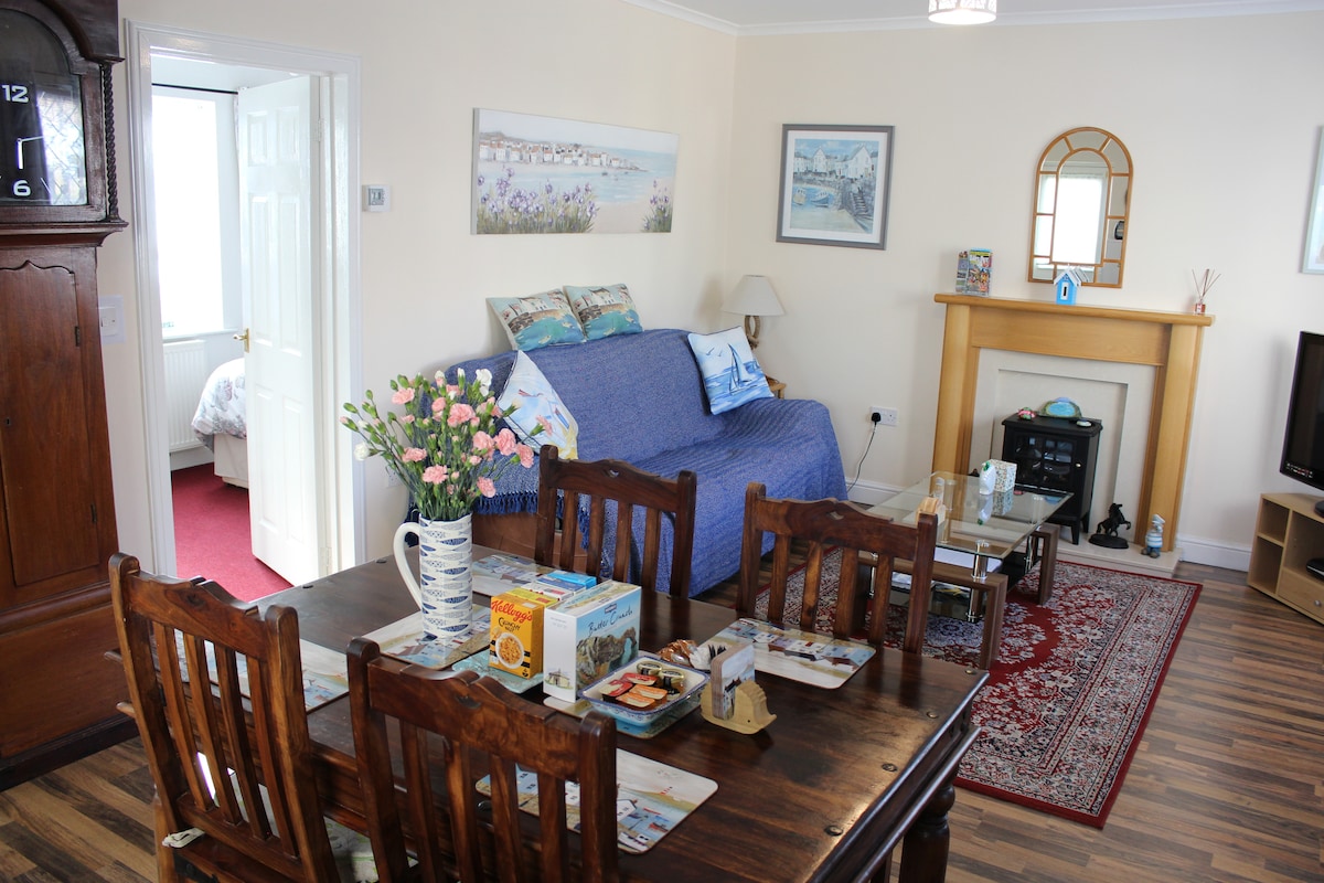 An open-plan kitchen living room features a dining table with wooden chairs and a cozy seating area. A blue sofa is situated against the wall, complemented by decorative pillows. A fireplace and artwork are visible, adding warmth to the space.